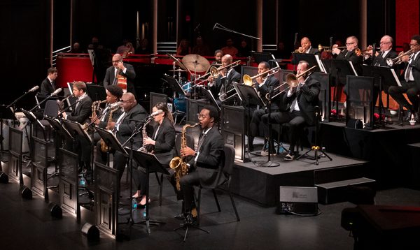 An orchestra plays on stage, in front of a red background