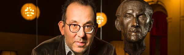 A man wearing glasses, a shirt and a corduroy jacket leans on top of a grand piano looking into the camera. Behind him is a bronze bust of theatre director Walter Felsenstein.