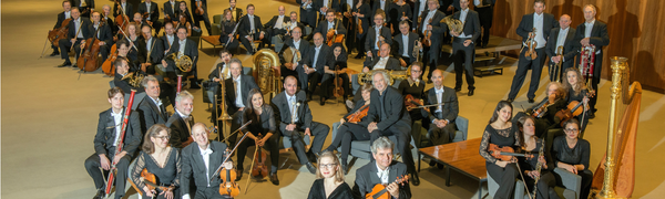 Deutsche Oper Berlin sit scattered around a large room. They are all holding their instruments and looking at the camera.
