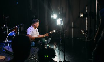 A man sits in a studio surrounded by cameras and lights playing a traditional Persian guitar-like instrument