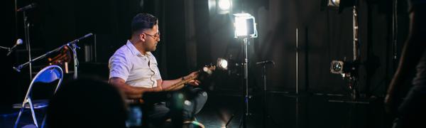 A man sits in a studio surrounded by cameras and lights playing a traditional Persian guitar-like instrument