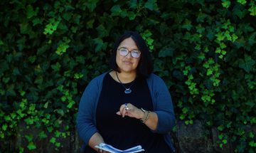 A woman sits in front of a wall of ivy reading from a book open in front of her