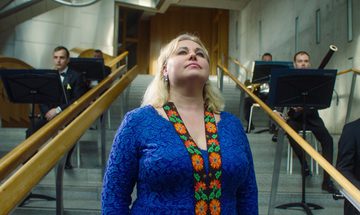 A blonde woman in a blue dress with an intricate flowered band stands on a set of steps in front of musicians