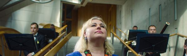 A blonde woman in a blue dress with an intricate flowered band stands on a set of steps in front of musicians