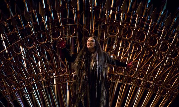 A woman dressed in all black stands in front of a fence made of spears with a terrifying look on her face