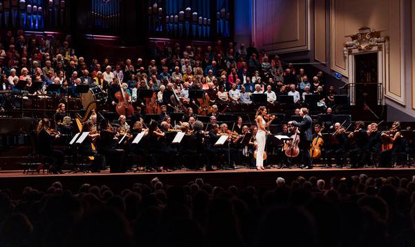 A solo violinist in a white jumpsuit stands onstage next to a conductor, with rows of seated orchestra members and a full organ gallery visible behind them.