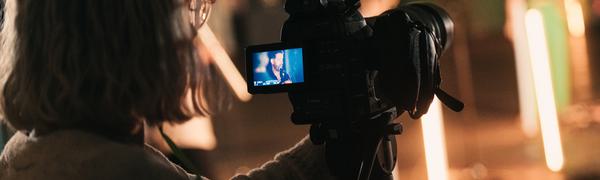 A woman stands behind a camera, filming a performer in front of her.
