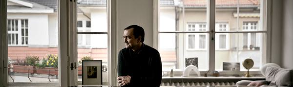 A man stands in front of large windows leaning against the top of a grand piano