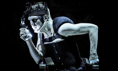 A man sits in a crown and a corset holding a microphone