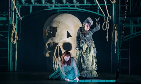 A woman kneels on the ground looking into a hole whilst a witch stands looming over her, with a giant skull visible behind them