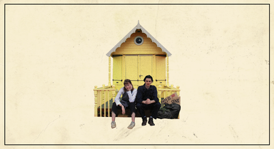 A young man and woman sit looking bored on the steps in front of a small yellow house