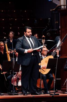 A man with dark hair in a black suit stands singing from a folder in front of him