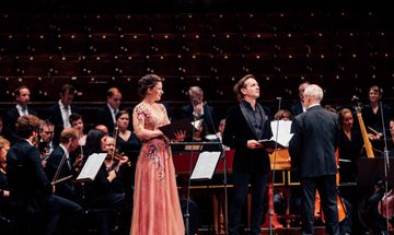 A woman in a pink dress and a man in a black suit stand singing at the front of a stage in front of a full orchestra