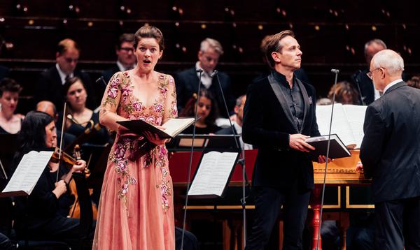 A woman in a pink dress and a man in a black suit stand in front of a full orchestra singing