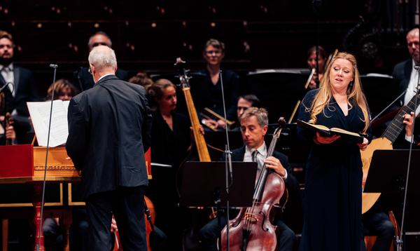 A woman with long blonde hair in a black dress stands singing in front of a full orchestra