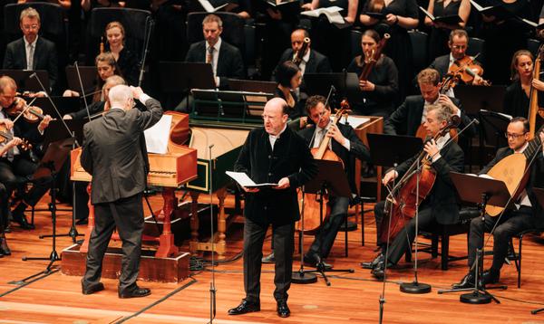 A man in a black jacket stands in front of a full orchestra singing with an angry expression on his face