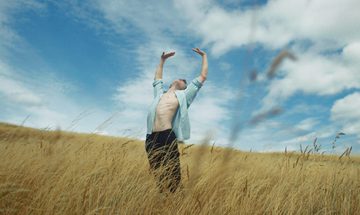 A man in an open blue shirt stretches his arms above him in a field of golden corn against a blue sky