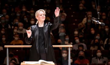 A woman dressed in all black stands with a full audience behind her, with one hand in the air and a conductor's baton in her other hand.