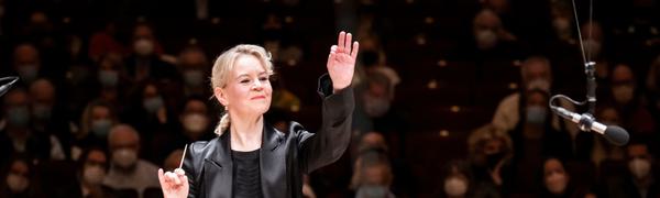 A woman dressed in all black stands with a full audience behind her, with one hand in the air and a conductor's baton in her other hand.