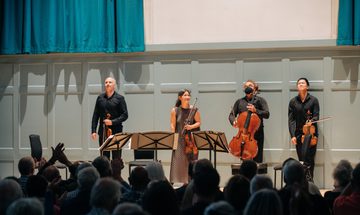Four string players stand in front of an applauding audience against a light blue wall