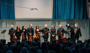 A group of musicians dressed in black holding string instruments stand on stage looking out at an applauding crowd