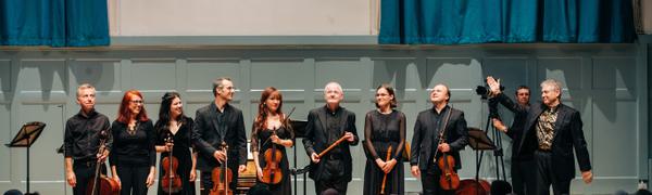 A group of musicians dressed in black holding string instruments stand on stage looking out at an applauding crowd