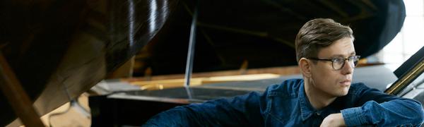 A man sits in between two grand pianos with one arm resting under the lid of one of the pianos.