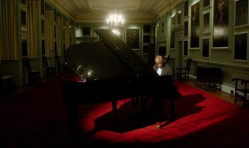 A Black man in a black suit sits at a grand piano under a spotlight, surrounded by portraits on the walls
