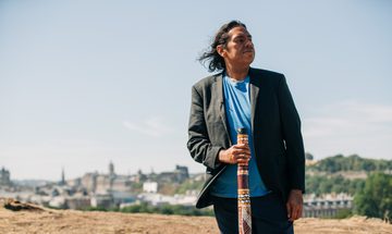 An aboriginal man in a bright blue tshirt and black blazer stands holding a didgeridoo with a view of Edinburgh behind him