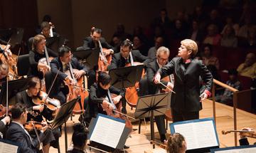 A conductor onstage begins to raise her arms, with the string section of the orchestra visible in front of and around her and the seated audience behind.