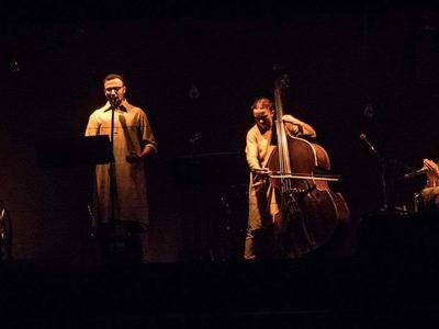 Four musicians spotlit onstage, each dressed in neutral colours and standing or seated behind a music stand and microphone.