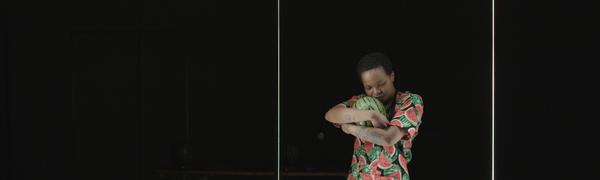 a black stage with a solitary figure, dressed in a watermelon print dress and cradling a watermelon, standing behind a white frame