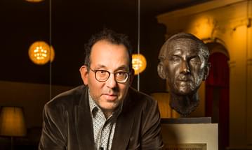 A man wearing glasses, a shirt and a corduroy jacket leans on top of a grand piano looking into the camera. Behind him is a bronze bust of theatre director Walter Felsenstein.