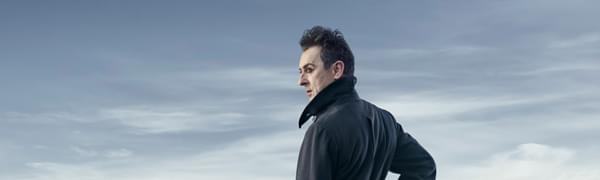 Alan Cumming pictured in profile standing on top of a snowy mountain, wearing a long black coat with the collar turned up.
