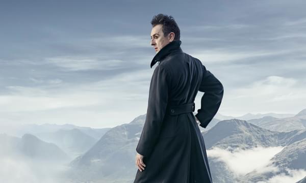 Alan Cumming pictured in profile standing on top of a snowy mountain, wearing a long black coat with the collar turned up.