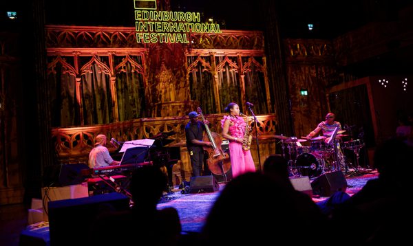 A woman in a ruffled pink outfit plays the saxophone alongside a pianist, a double-bassist, and a drummer. A yellow sign overhead reads Edinburgh International Festival.