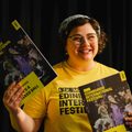 A smiling white woman wearing a yellow Edinburgh International Festival t-shirt holds up programmes for A Masked Ball
