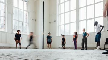 A group of people in exercise clothes stand spaced out in the corner of a white studio space with large warehouse windows.