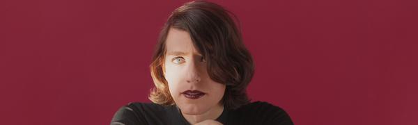 A woman with dark shoulder-length hair sits at a table with her arms folded against a maroon background.