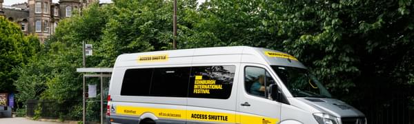 A silver bus with Edinburgh International Festival branding is parked at a bus stop in front of trees and buildings on a quiet road.