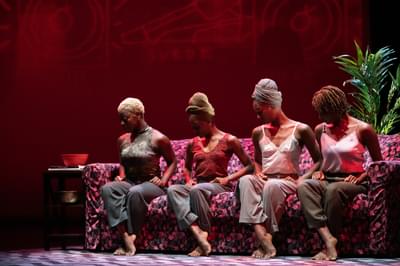 A row of four woman sit on a sofa, dressed in casual clothes and head wraps, in matching poses with their heads bowed, ankles crossed and toes pointed.