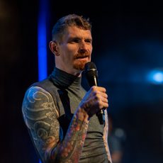 A man with a full sleeve of arm tattoos and a trim beard speaks into a microphone.