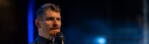 A man with a full sleeve of arm tattoos and a trim beard speaks into a microphone.