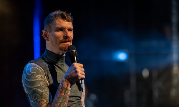 A man with a full sleeve of arm tattoos and a trim beard speaks into a microphone.