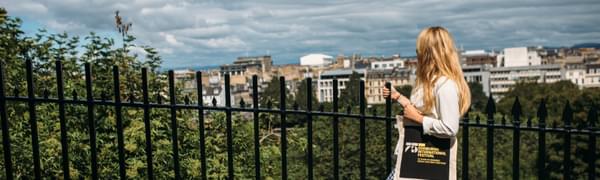 A woman with long blonde hair stands with her back to the camera, leaning against railings and looking out over the Edinburgh skyline, holding a tote bag featuring the logo of the 75th Edinburgh International Festival.