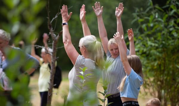 A group of women standing outside i a leafy park stretch their arms up towards the sky.
