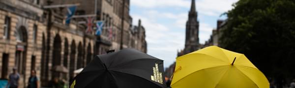 A view of an Edinburgh street with two Edinburgh International Festival umbrellas in the foreground, one yellow, one black.