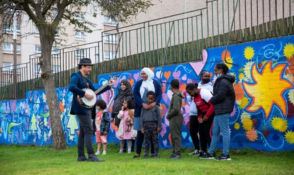 A man plays a banjo to a group of children and their mothers gathered in front of a colourful mural.