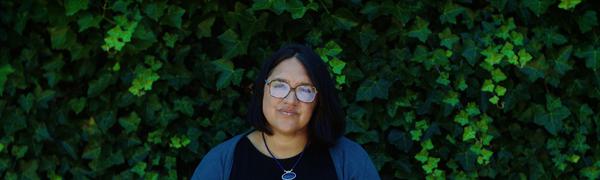 A woman sits in front of a wall of ivy reading from a book open in front of her