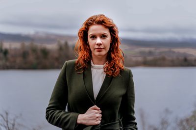 Woman with red wavy hair, a white shirt and a green blazer with a blurry background of a loch and hills